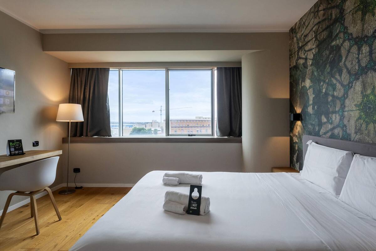 A hotel room with a large bed, a desk, a chair, a window with curtains, and a city view.