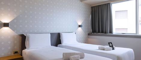 A hotel room with two beds, a wooden nightstand, a window with curtains, and wall-mounted lights.