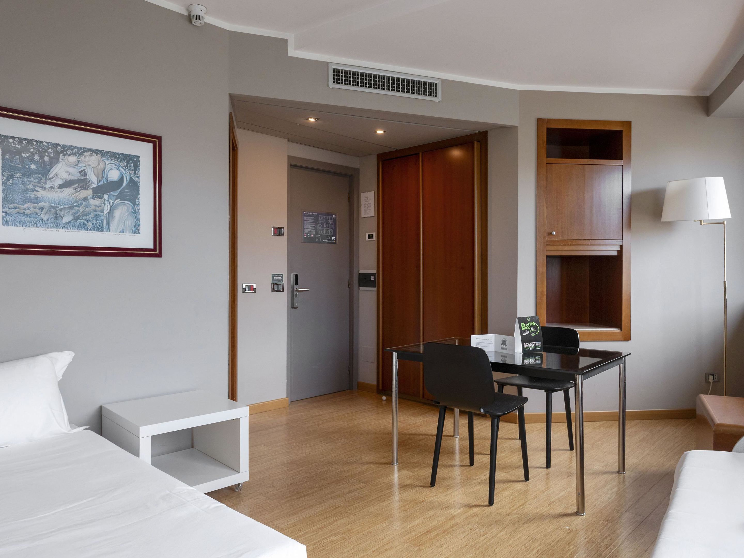 A hotel room with a bed, a desk with a chair, a framed picture on the wall, a wooden cabinet, and a lamp.