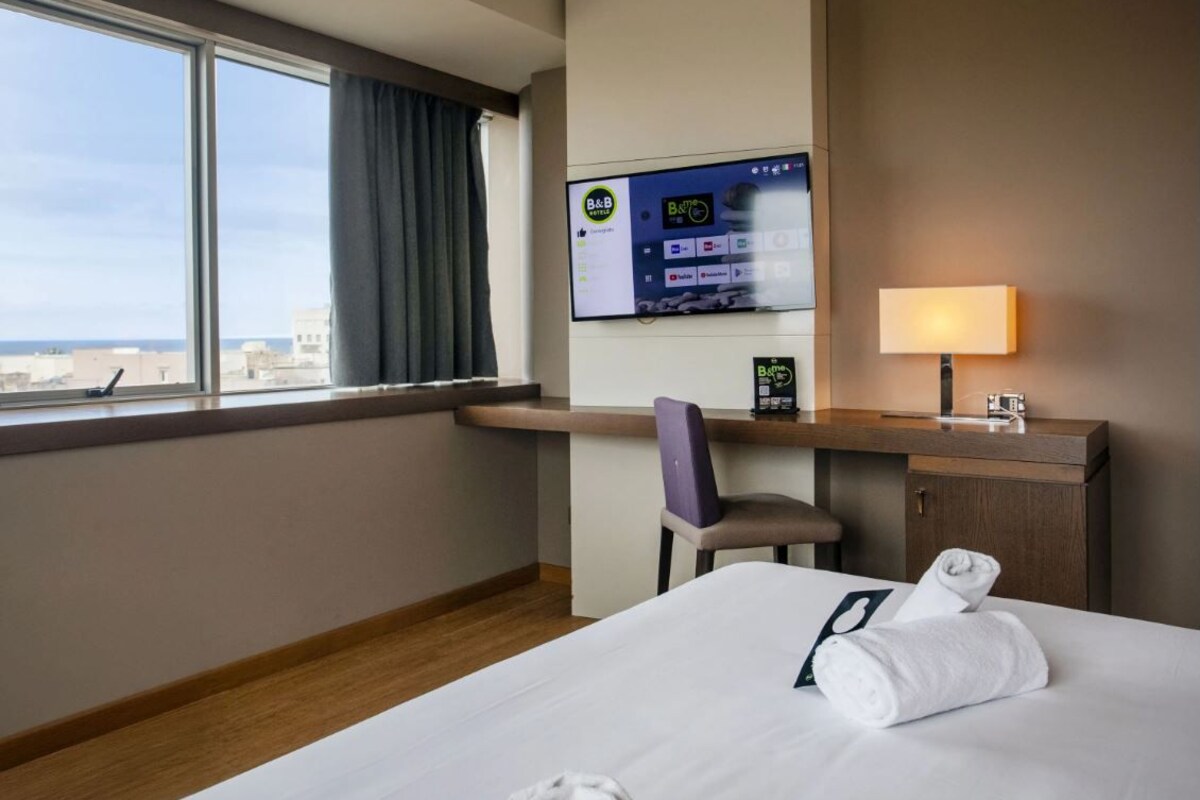 A hotel room with a bed, a desk with a computer monitor, a chair, a lamp, and a window with a view of the city.