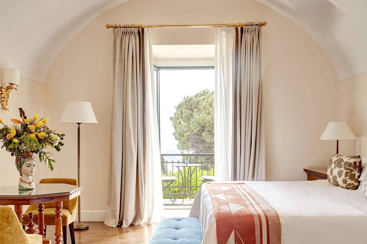 Un dormitorio con una cama grande, suelo de madera, balcón con cortinas y un candelabro.