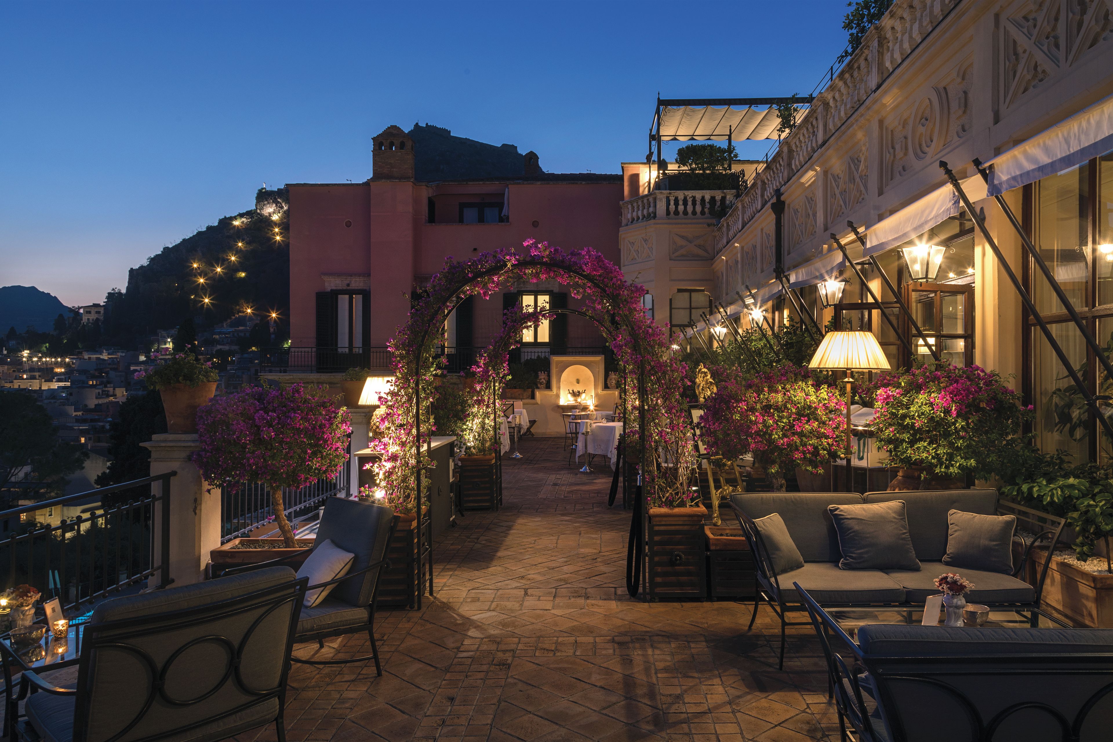 An outdoor seating area with a covered archway, tables, chairs, and potted plants.