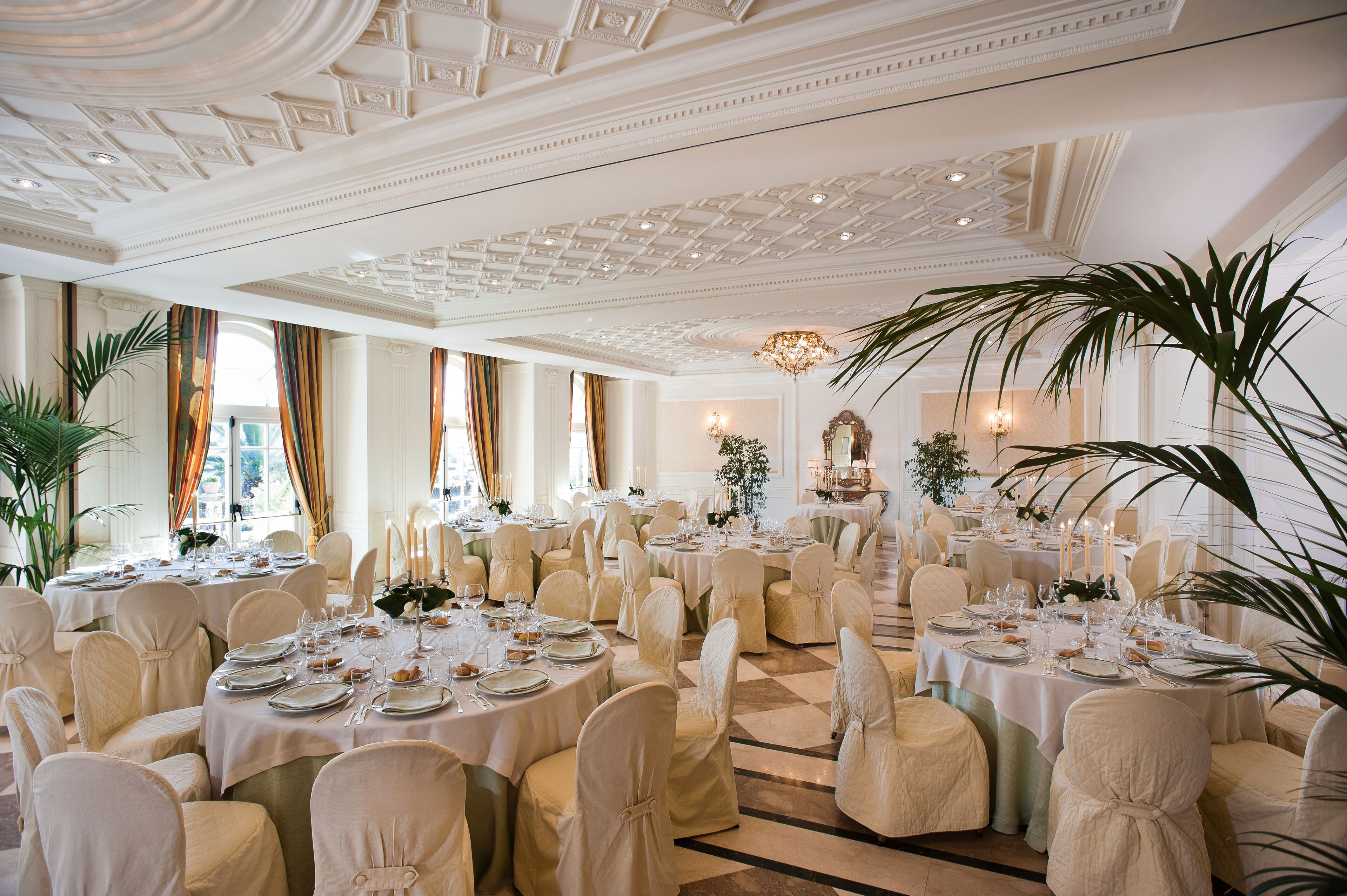 A banquet hall with round tables set for dining, white drapery, and a classic ceiling design.