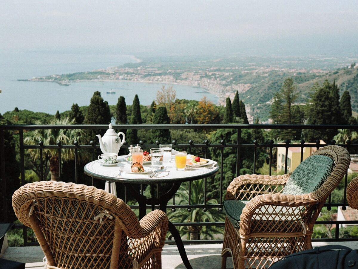 Uma varanda com cadeiras de vime, uma mesa redonda com itens de café da manhã e uma vista panorâmica de uma cidade costeira.