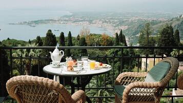 Uma varanda com cadeiras de vime, uma mesa redonda com itens de café da manhã e uma vista panorâmica de uma cidade costeira.