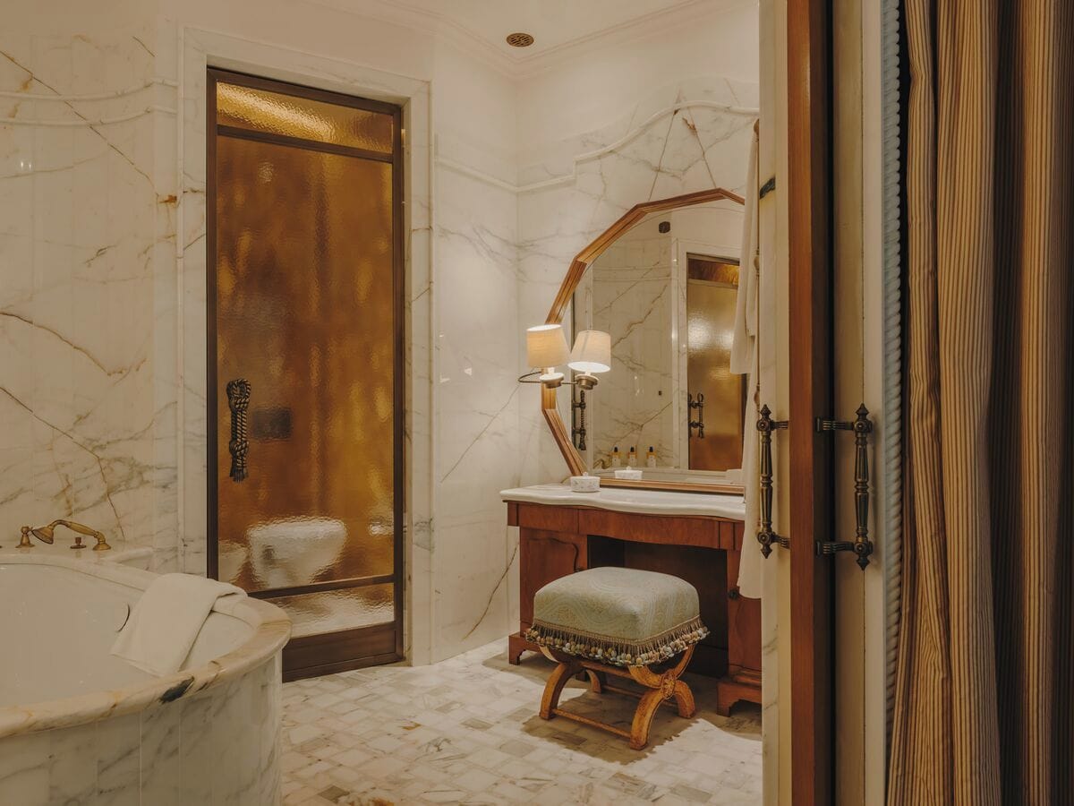 A bathroom with a marble floor, a bathtub, a wooden door, a vanity with a mirror, and a stool.