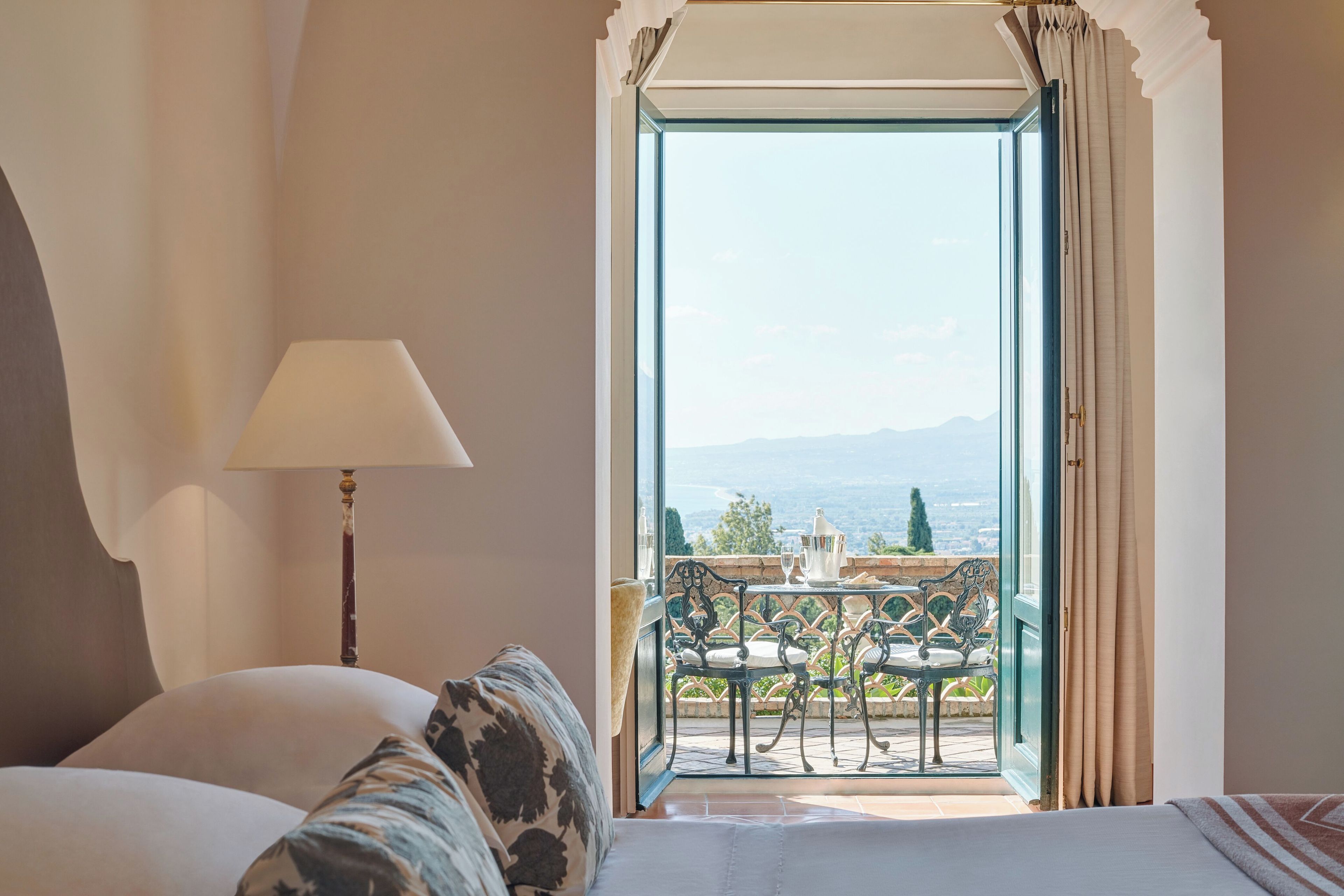 Un dormitorio con cama, una lámpara y vistas a un balcón exterior con mesa y sillas.