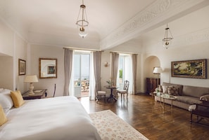 A hotel room with a large bed, a sofa, a dining table with chairs, and a balcony with curtains.