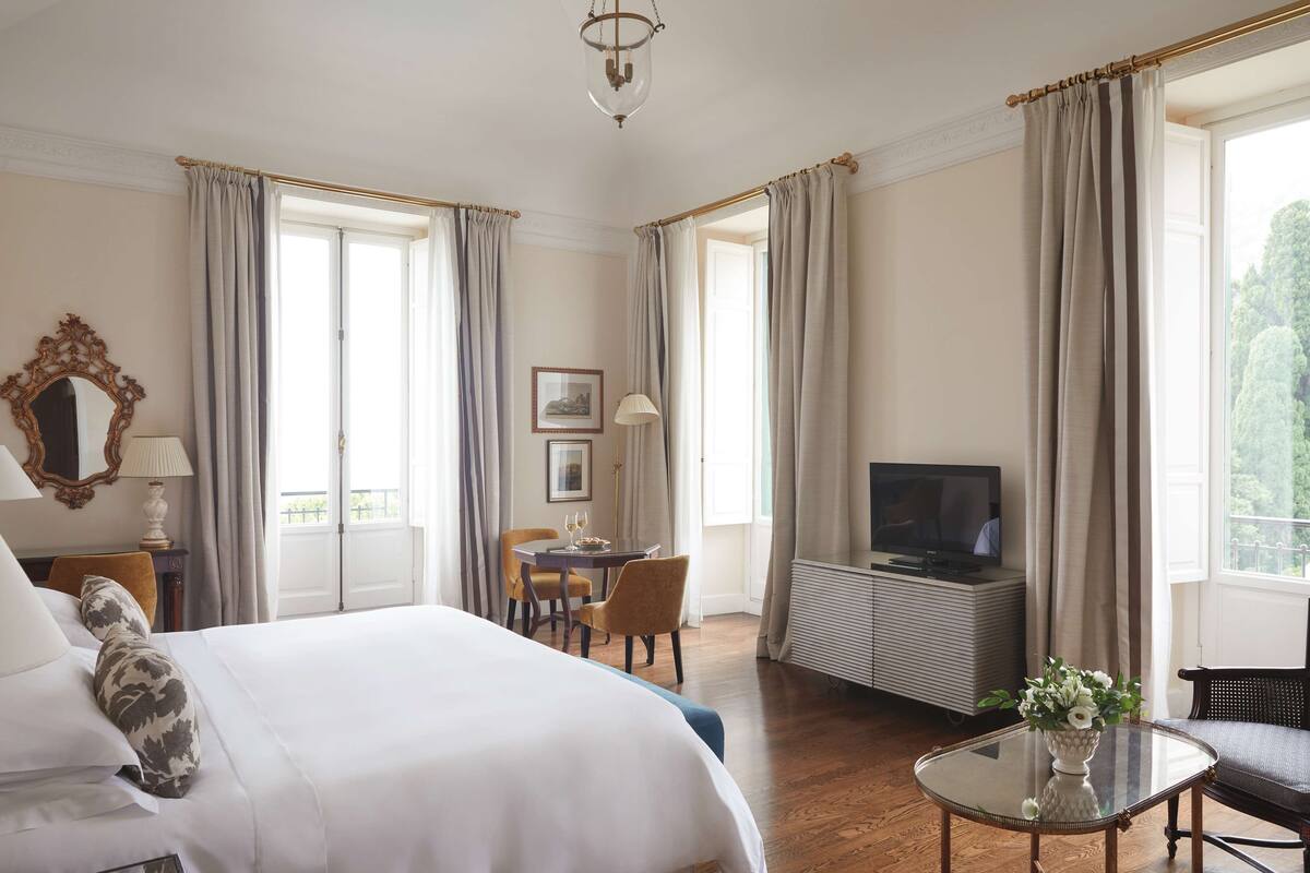 Habitación de hotel con una cama grande, televisión, mesa de comedor con sillas y una mesita con un florero.