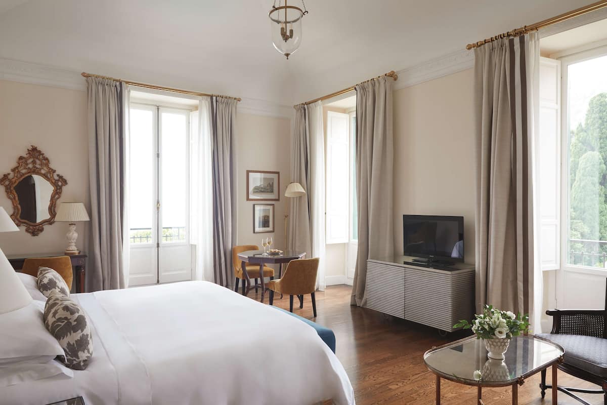 A hotel room with a large bed, a television, a dining table with chairs, and a small table with a vase.