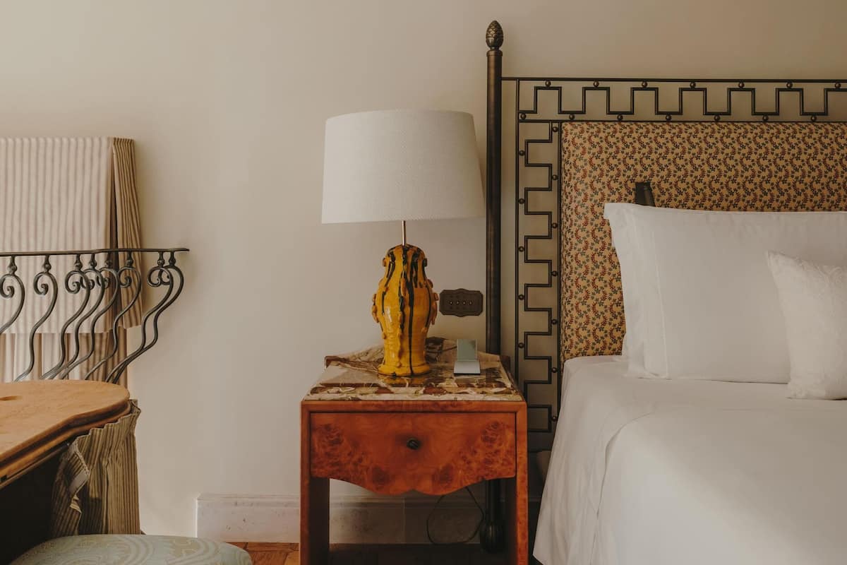 Una habitación de hotel con cama, mesita de noche con lámpara, una silla de madera y un cabecero estampado.
