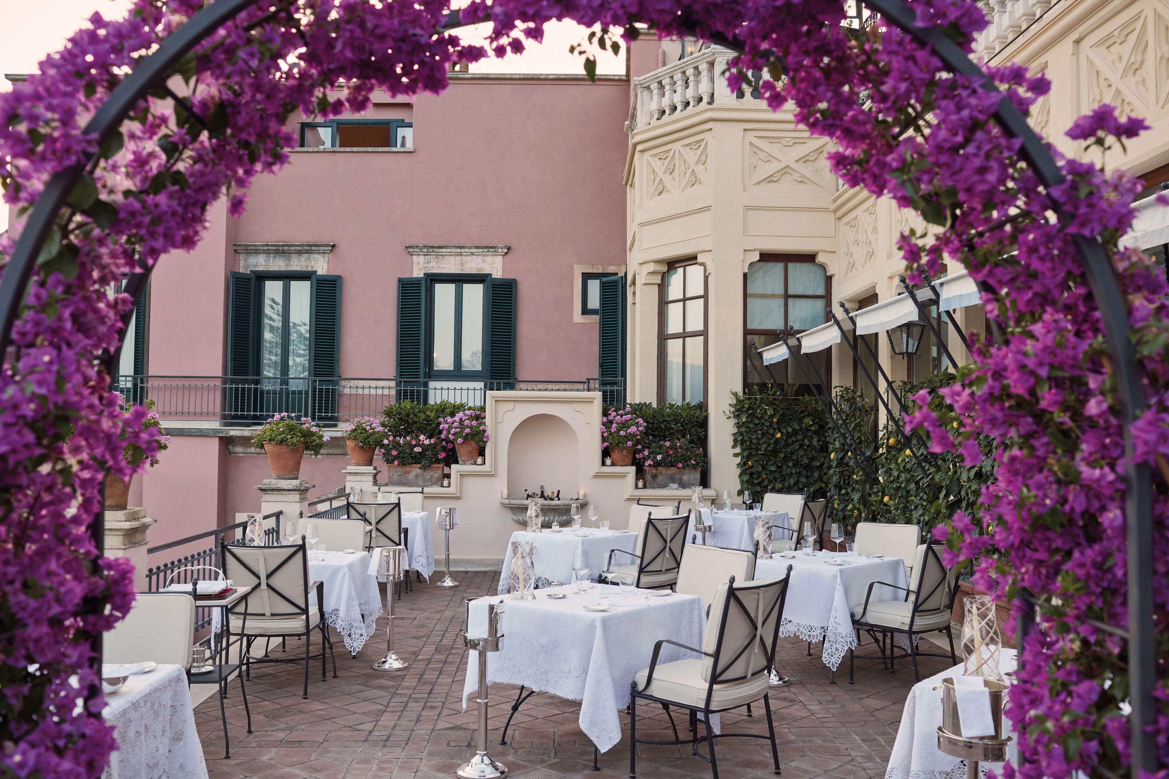 An outdoor patio with tables and chairs, surrounded by a pink building with green shutters and potted plants.