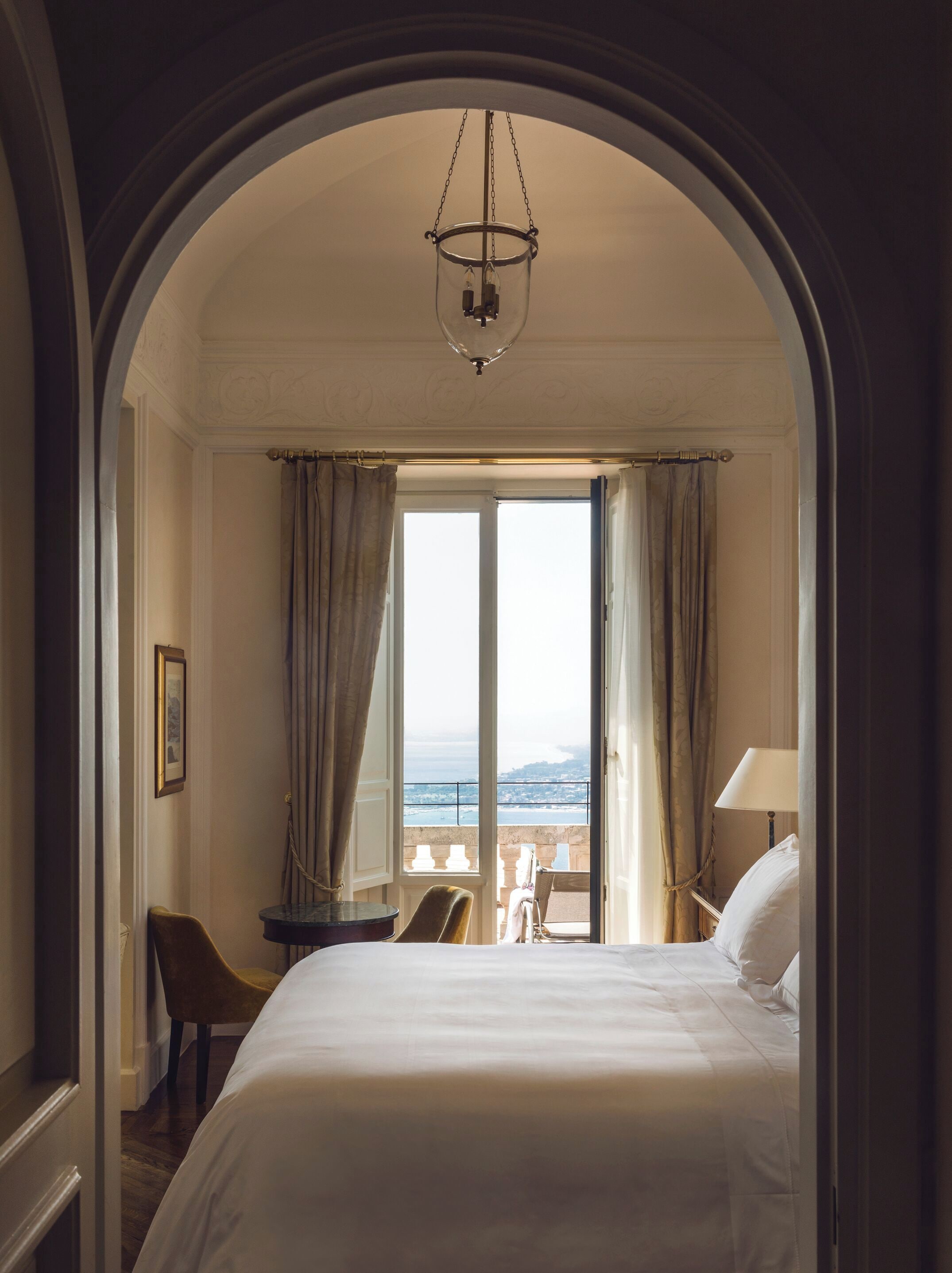 A hotel room with a bed, a chair, a desk, and a view of the sea through a large window.