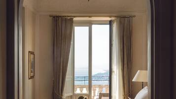 A hotel room with a bed, a chair, a desk, and a view of the sea through a large window.