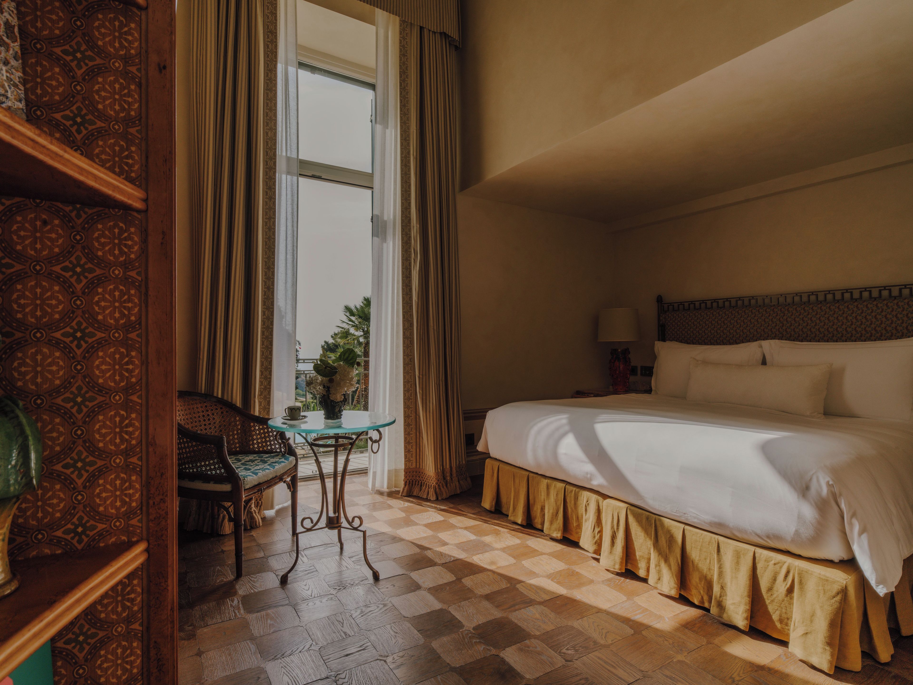 Una habitació d'hotel amb un llit gran, una taula petita amb un ram, una cadira i vista cap a l'exterior a través d'una finestra.