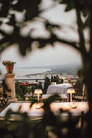 An outdoor dining area with tables and chairs, a potted plant, and a view of a body of water and distant landscape. - Grand Hotel Timeo, A Belmond Hotel, Taormina (Taormina)