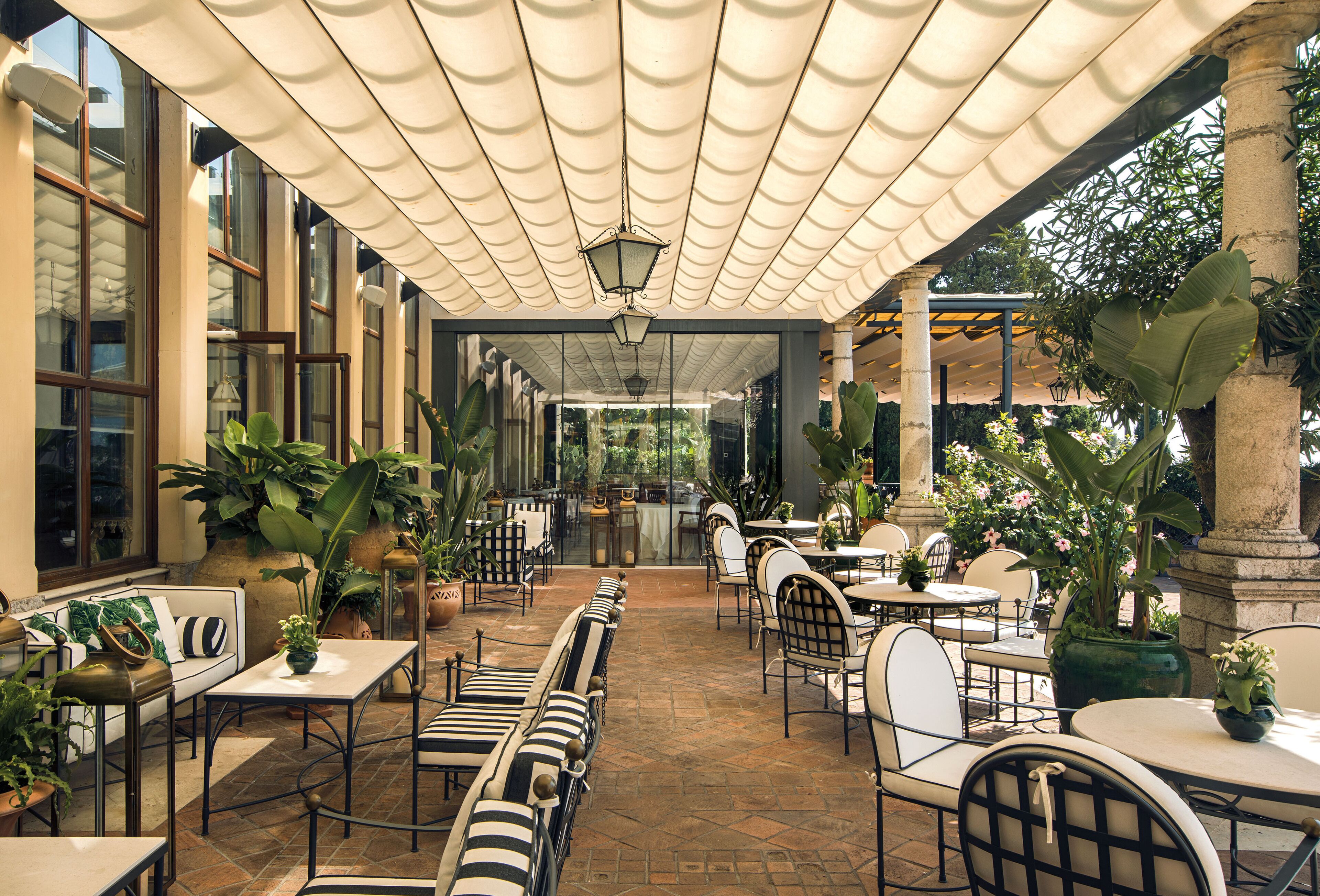 An outdoor patio area with tables and chairs, potted plants, and a covered walkway.