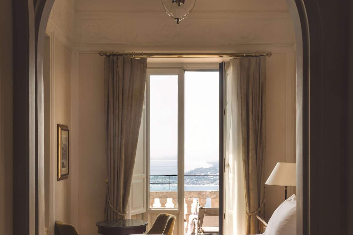 Un cuarto de hotel con una cama, una silla, un escritorio y vista al mar a través de un ventanal grande.
