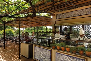 Un café al aire libre con zona de estar cubierta, una barra con un chef y plantas en macetas.