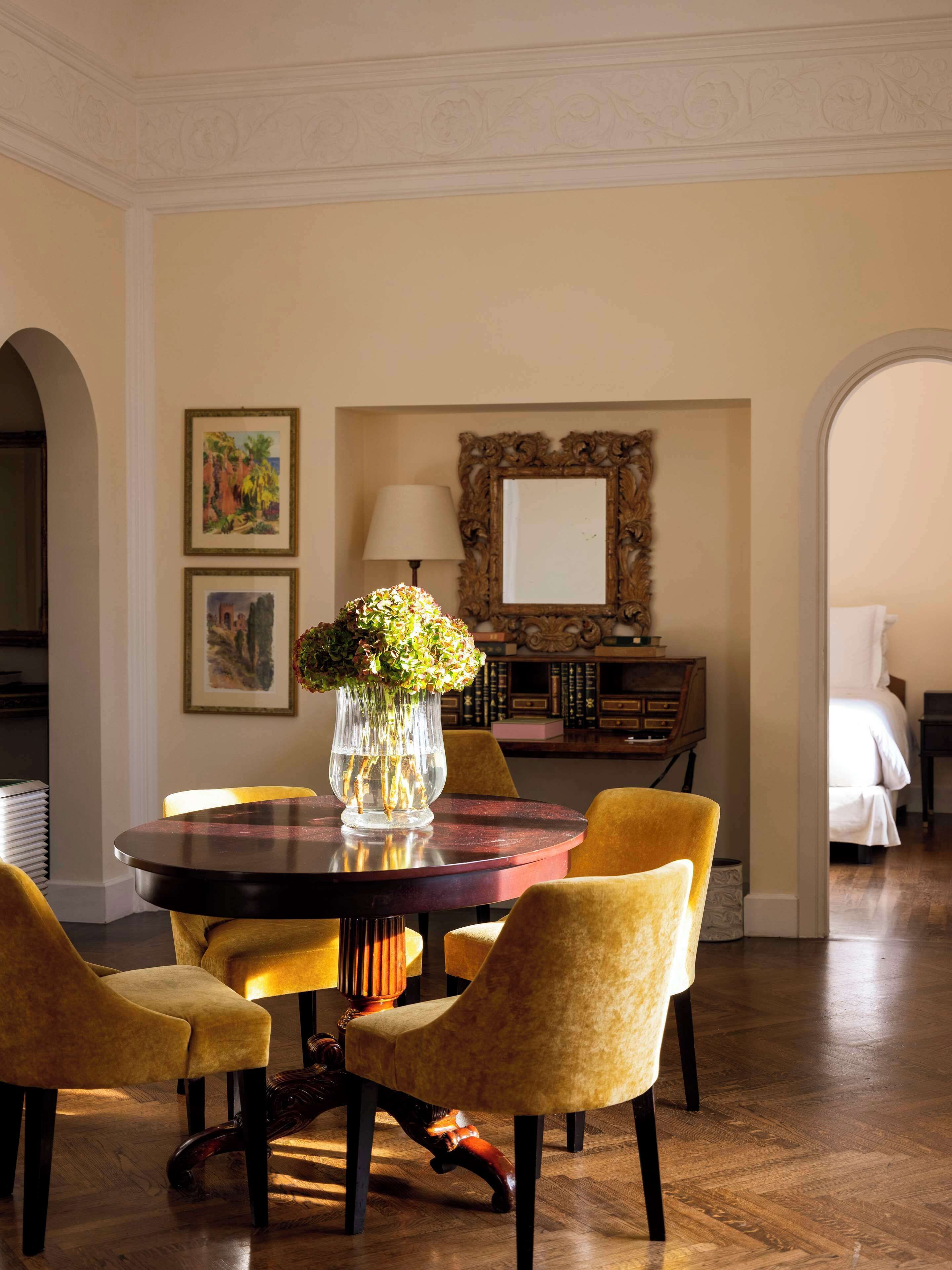 Un coin repas avec une table ronde, des chaises jaunes, un vase de fleurs, des œuvres d’art encadrées et un miroir.