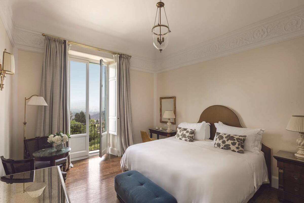 Un dormitorio con una cama grande, un escritorio con silla, una mesita con flores y vistas amplias a través de un ventanal.