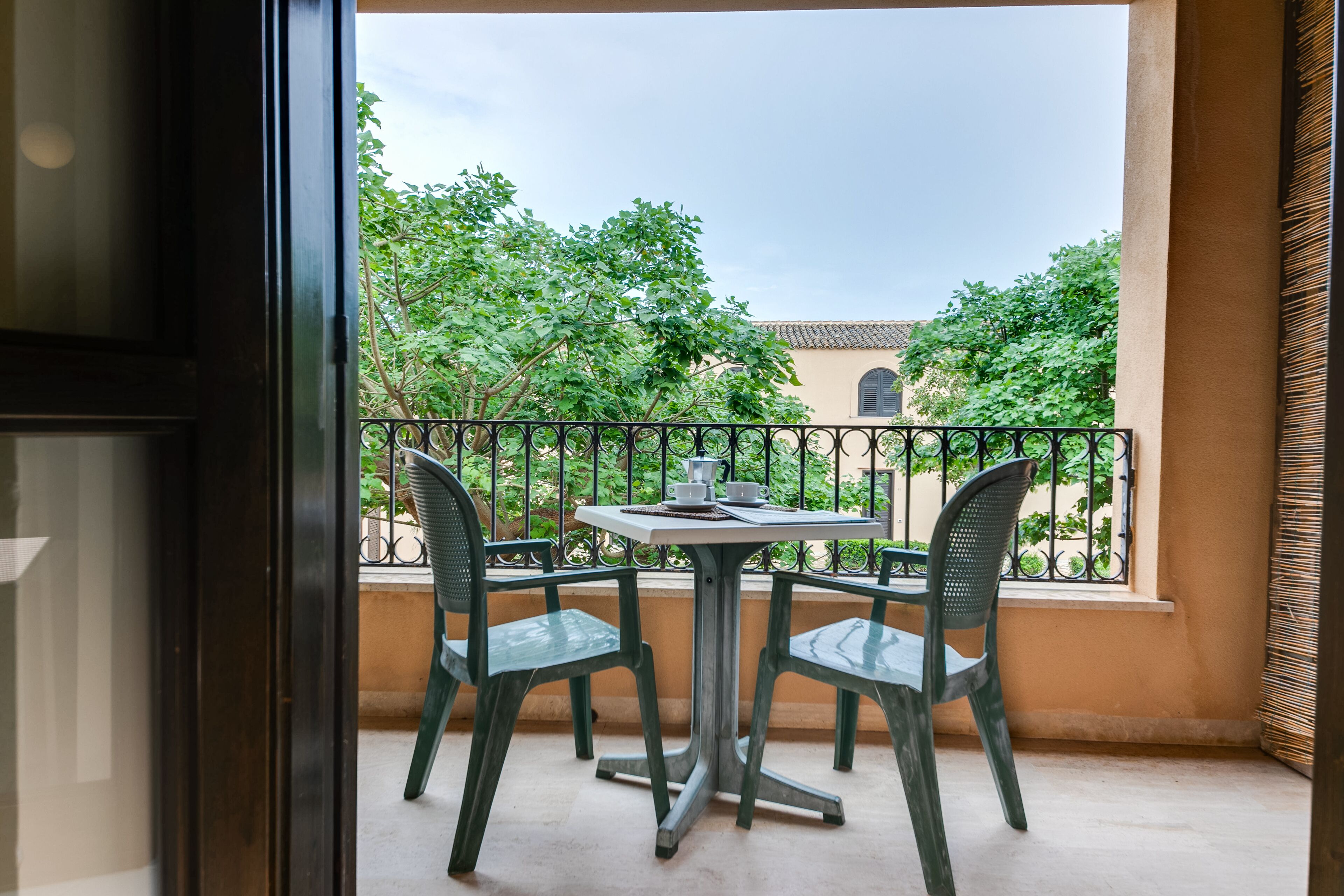 Uma varanda com mesa e cadeiras, vista para o verde e um edifício ao fundo.