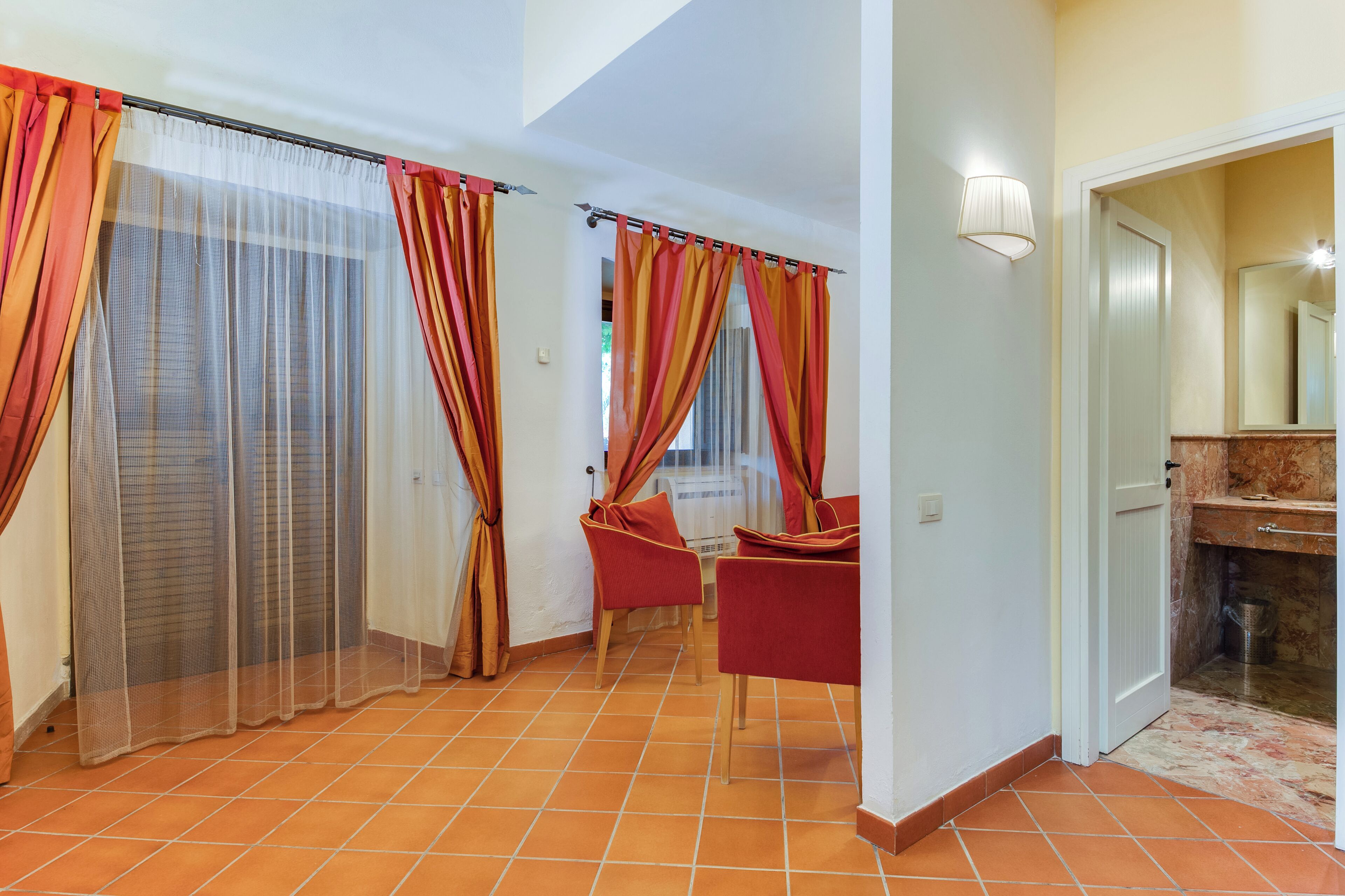 A hotel room with a tiled floor, a bed, a bathroom with a marble countertop, and red chairs in the corner.
