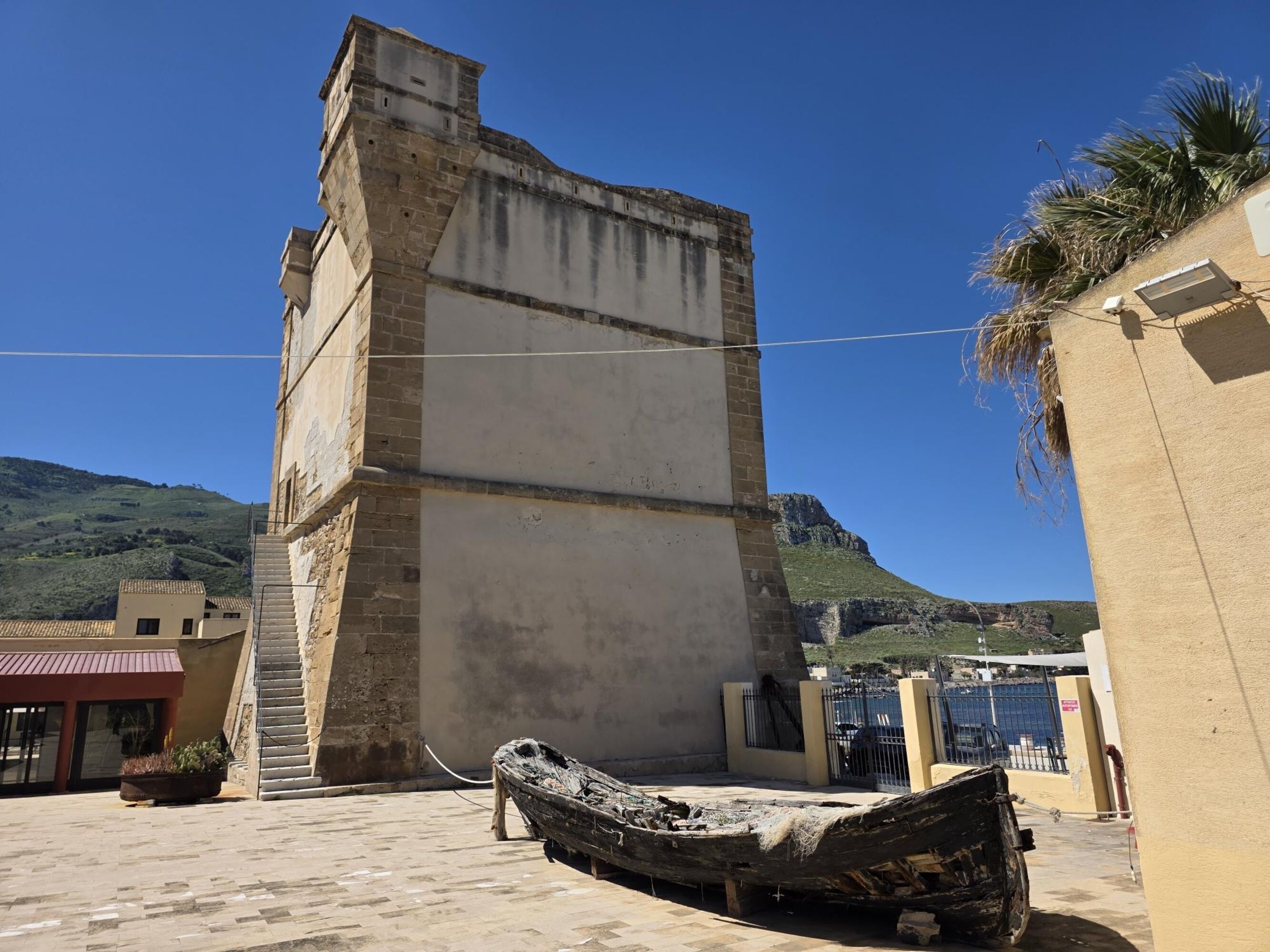 Edifici a la costa amb una torre alta, un vaixell i un paisatge de muntanyes al fons.