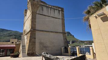 Edifici a la costa amb una torre alta, un vaixell i un paisatge de muntanyes al fons.