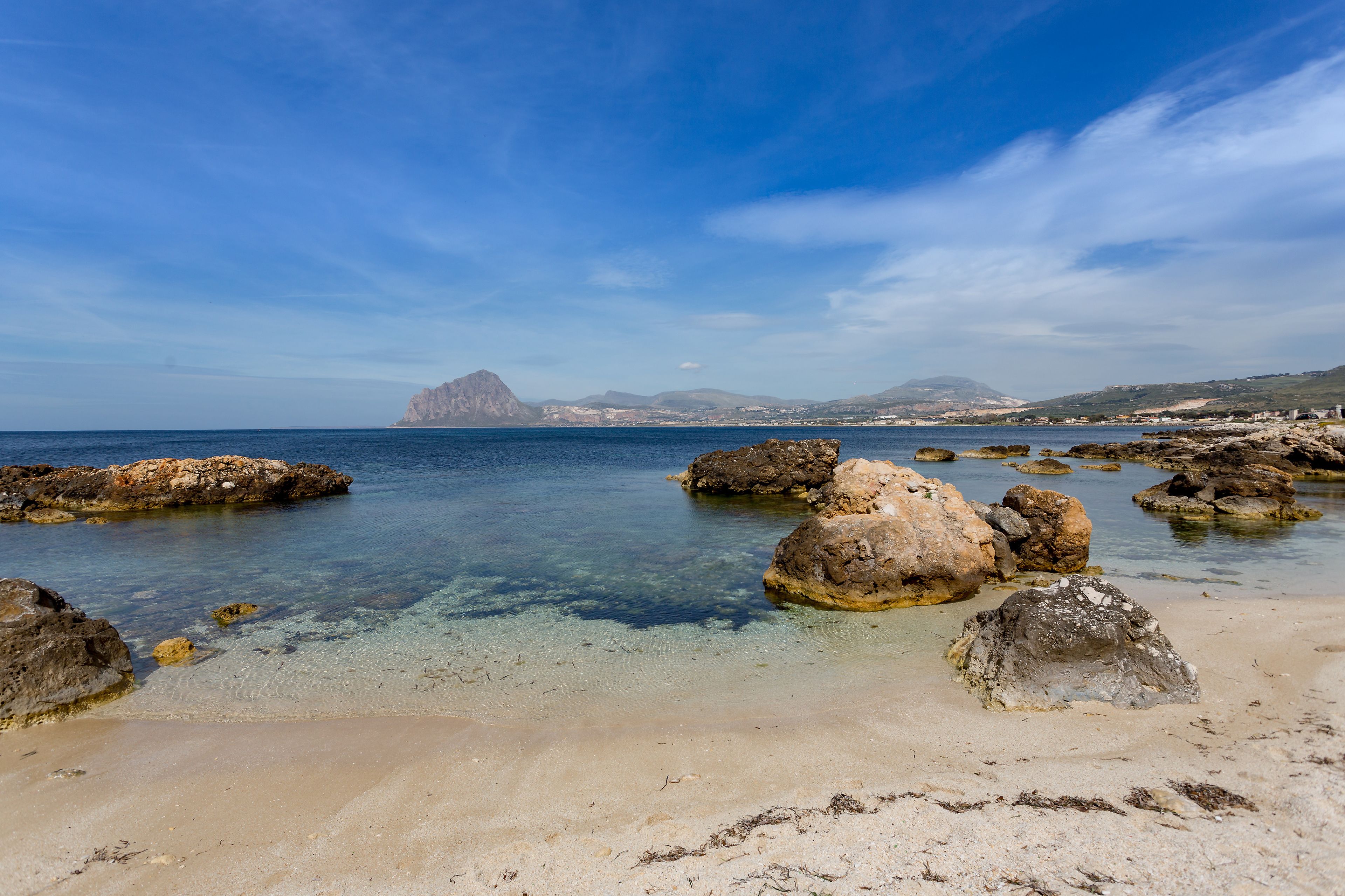 A coastal beach with clear water, large rocks, and distant mountains.