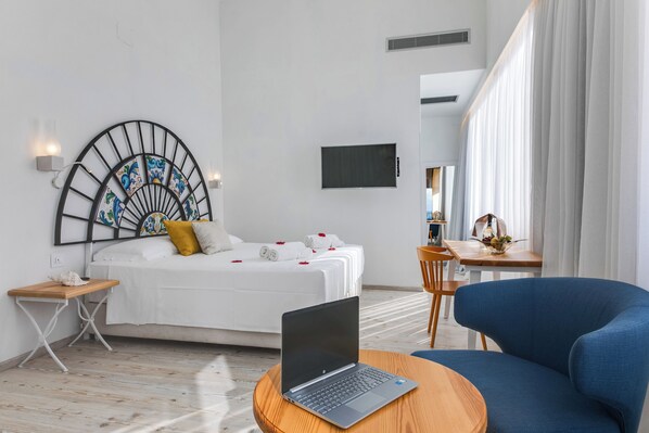 Habitación de hotel moderna con una cama, una laptop sobre una mesa redonda, un sillón azul, una mesita de noche de madera y un televisor montado en la pared.