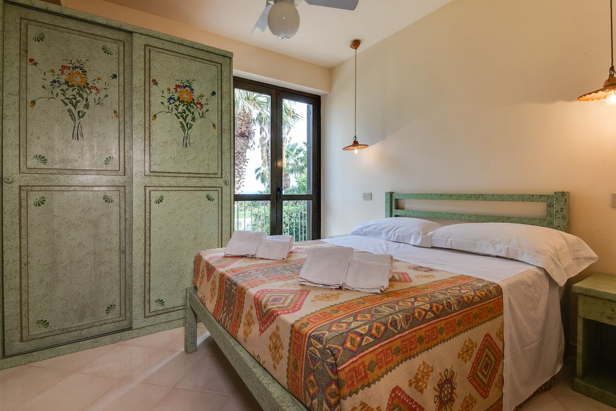 Une chambre à coucher avec un lit, une armoire verte et une fenêtre donnant sur des palmiers.