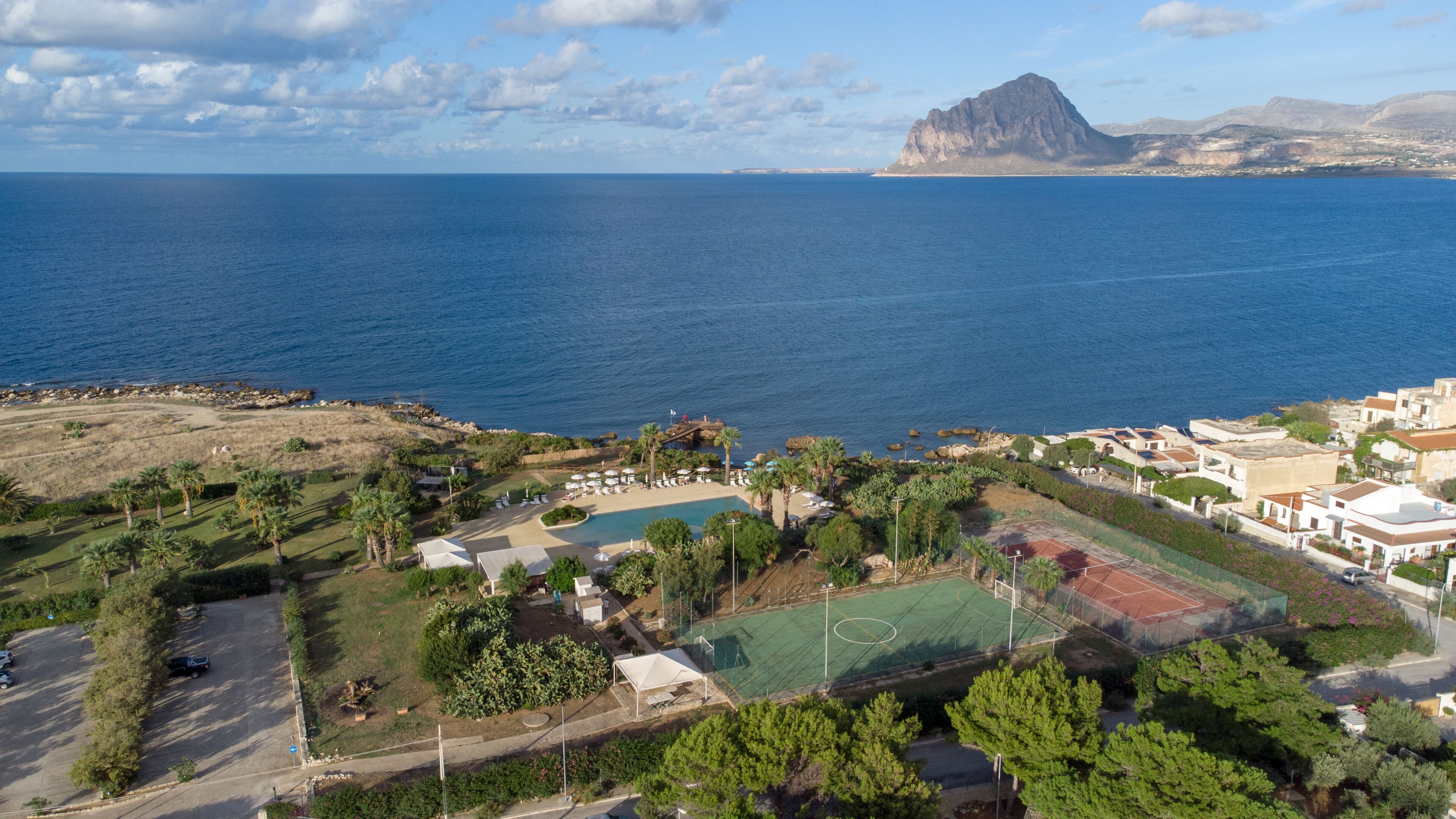 Un complejo turístico frente al mar con una piscina grande, canchas de tenis y una montaña rocosa al fondo.