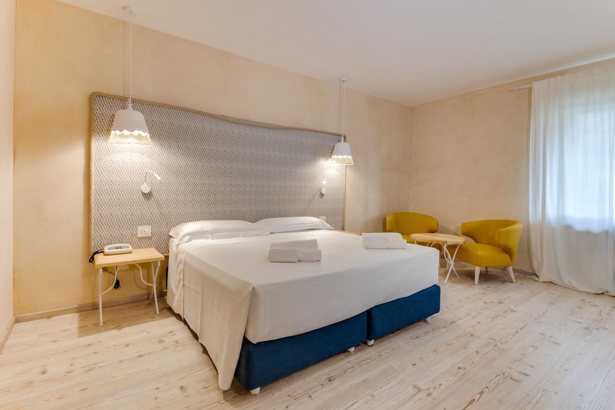 Une chambre d’hôtel moderne avec un grand lit, des tables de chevet, une petite table, deux chaises jaunes et une grande fenêtre avec des rideaux.