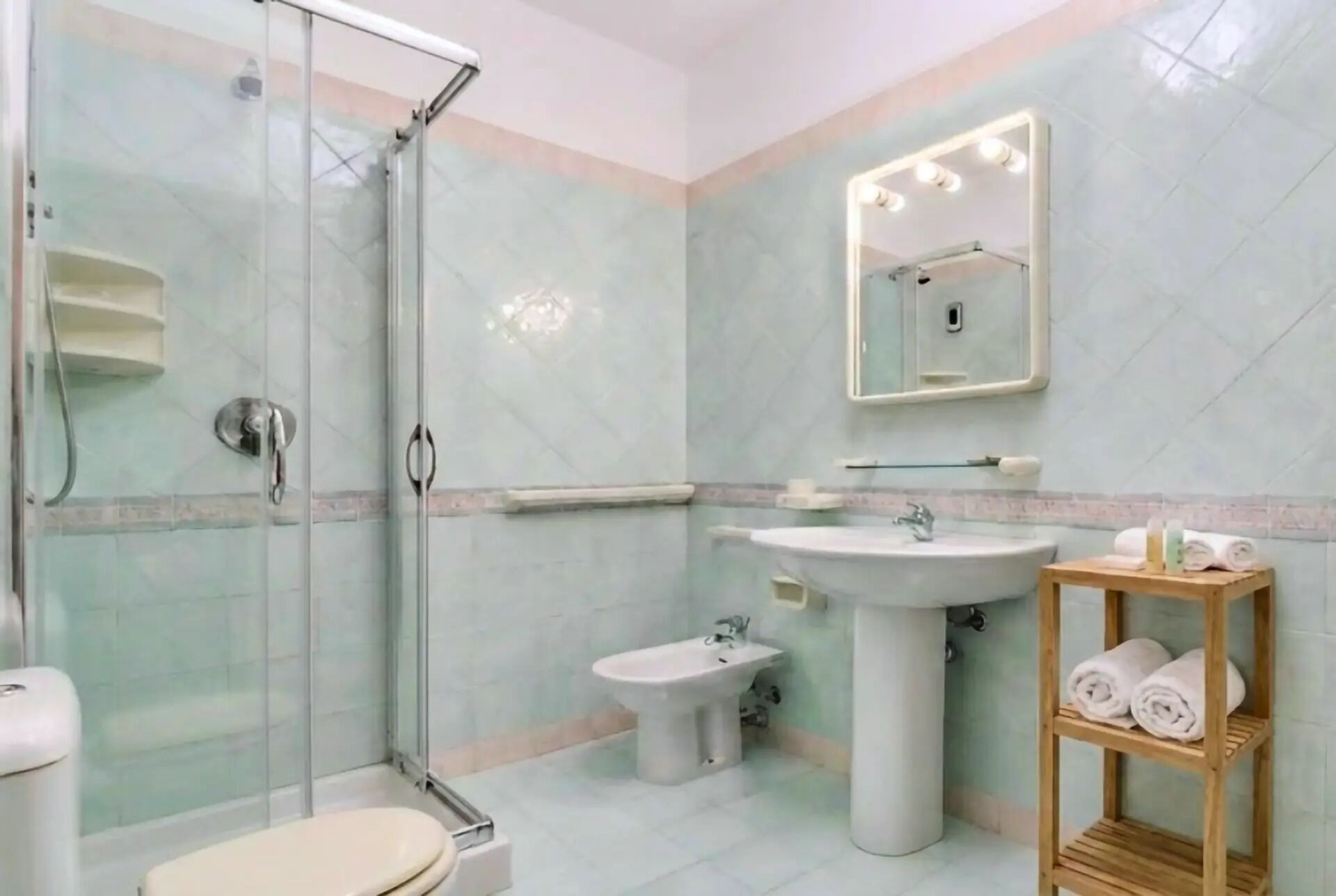 Une salle de bain dotée d’une douche à parois de verre, d’un lavabo sur colonne, d’un bidet et d’une étagère en bois où sont posées des serviettes.