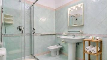 Une salle de bain dotée d’une douche à parois de verre, d’un lavabo sur colonne, d’un bidet et d’une étagère en bois où sont posées des serviettes.