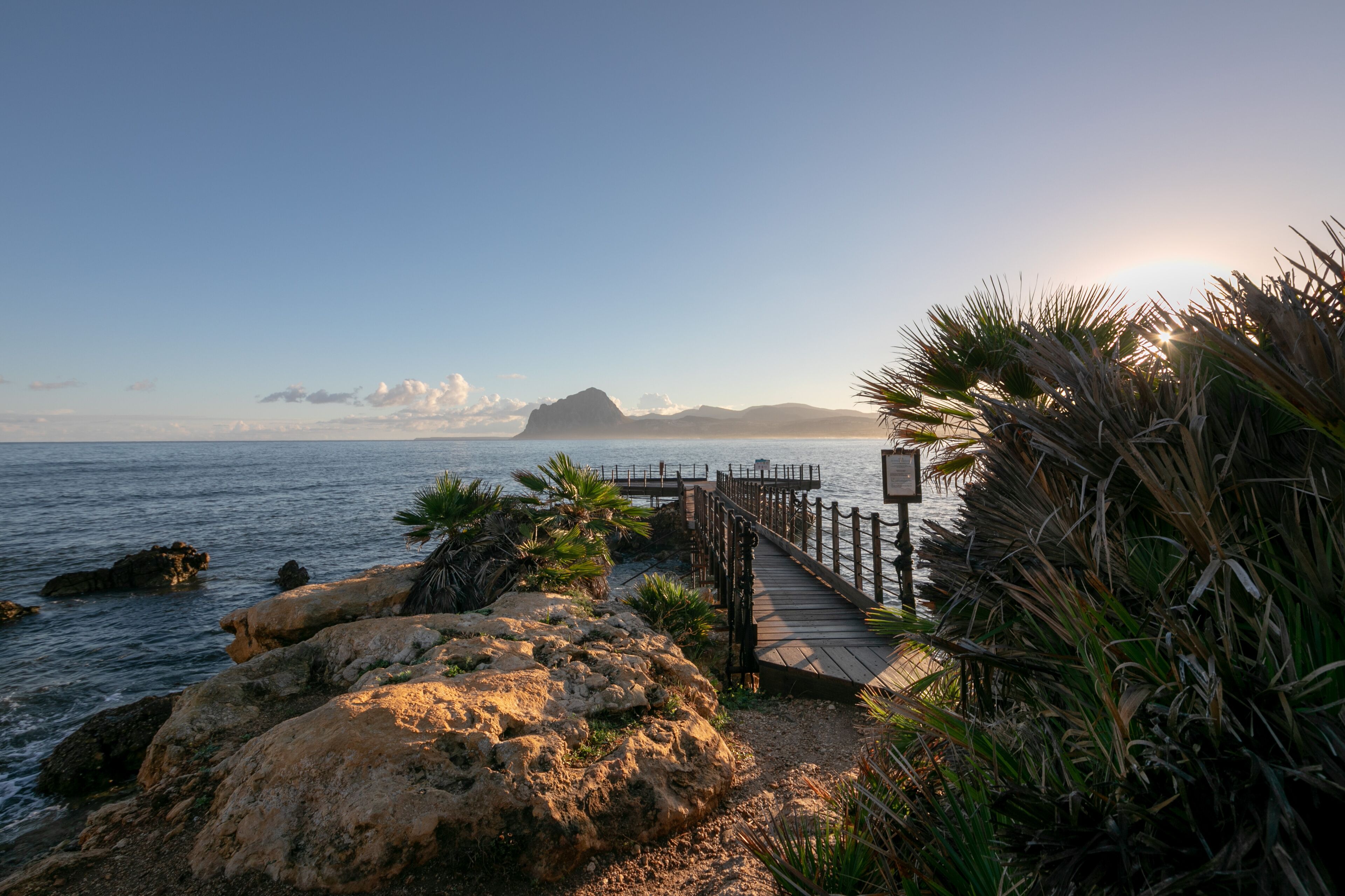 Une promenade en bord de mer avec une terrasse en bois, un terrain rocheux et des palmiers.