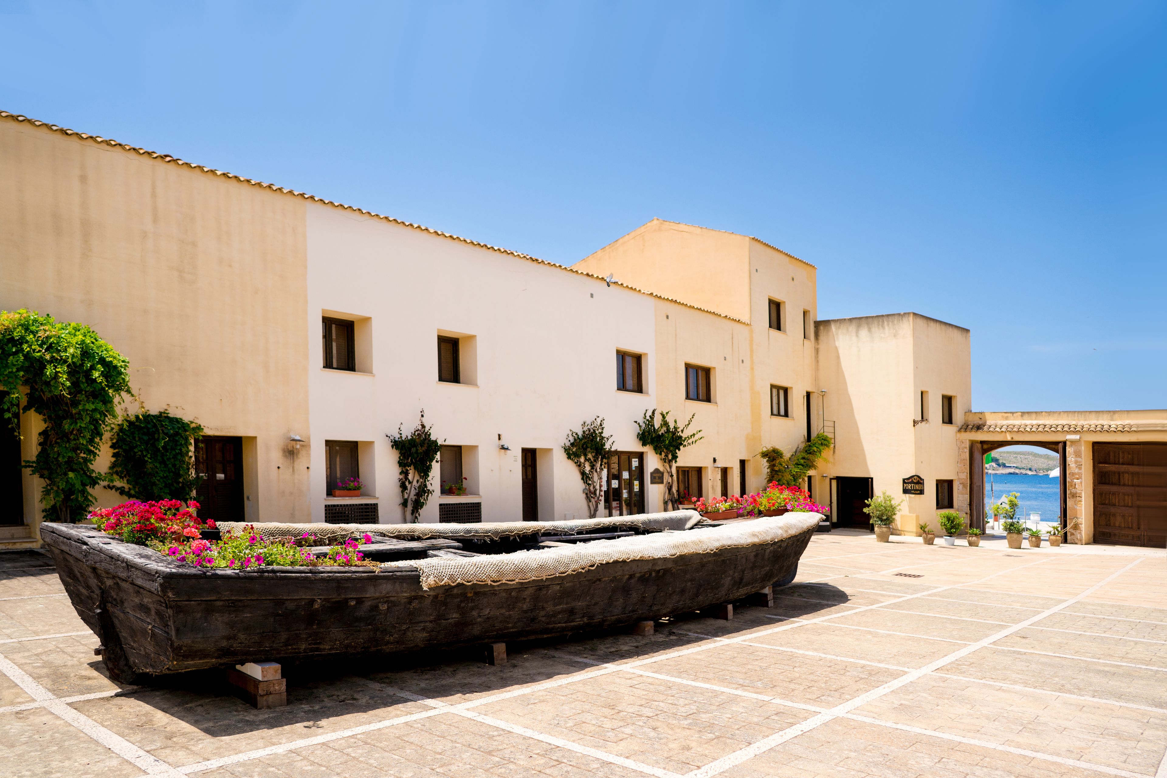 Un patio con una gran maceta con forma de barco llena de flores, suelo de baldosas y un edificio con varias ventanas y puertas.