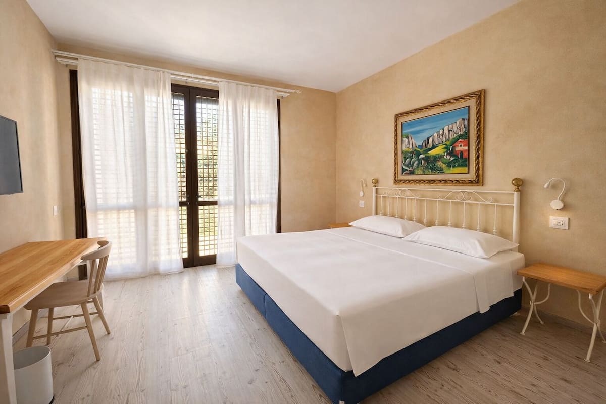 Une chambre à coucher avec un lit, un bureau, une chaise, une fenêtre avec des rideaux et un tableau au mur.
