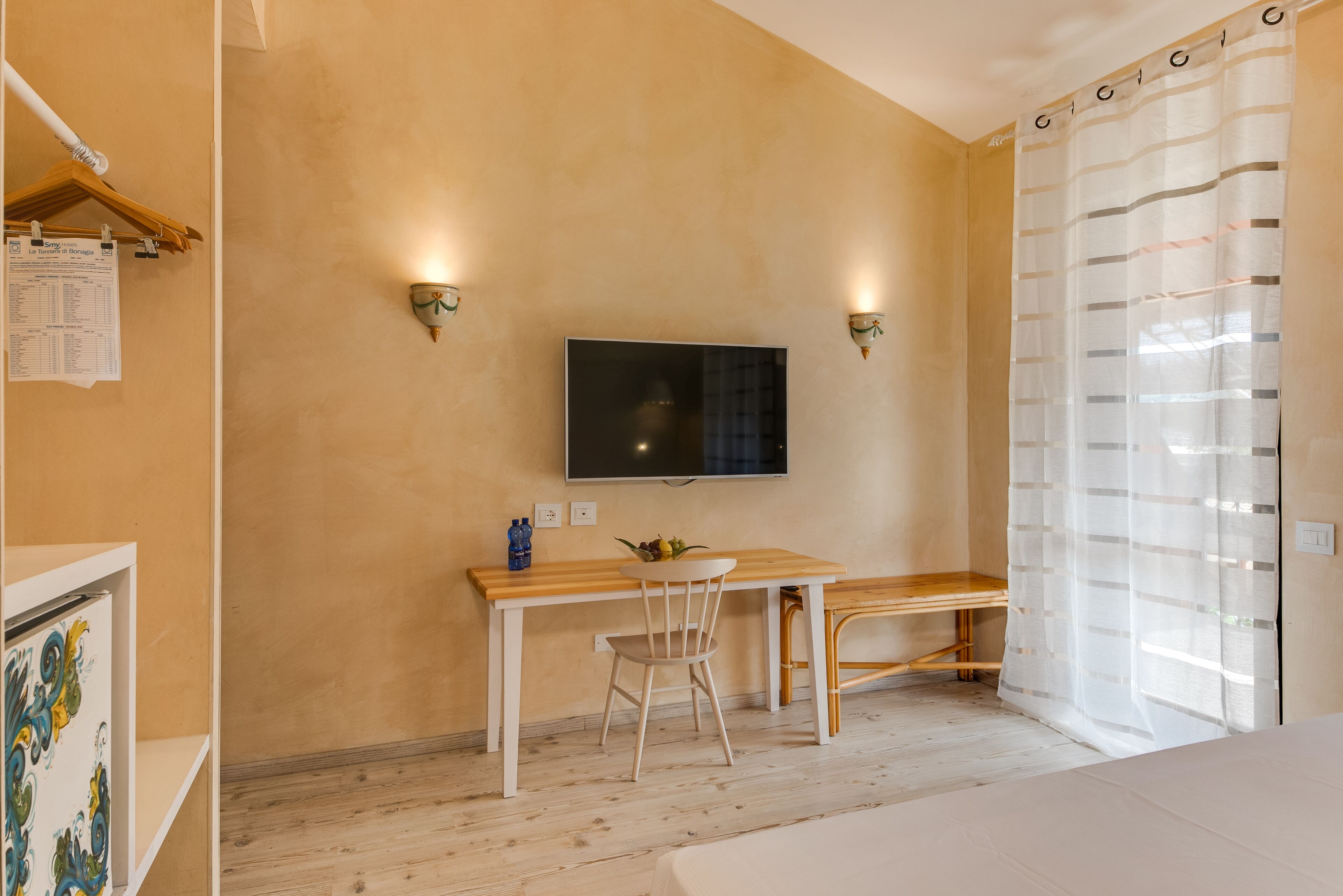 Ein Zimmer mit einem Holztisch, einem Stuhl, einem an der Wand montierten Fernseher und einem Bett mit weißer Bettdecke.