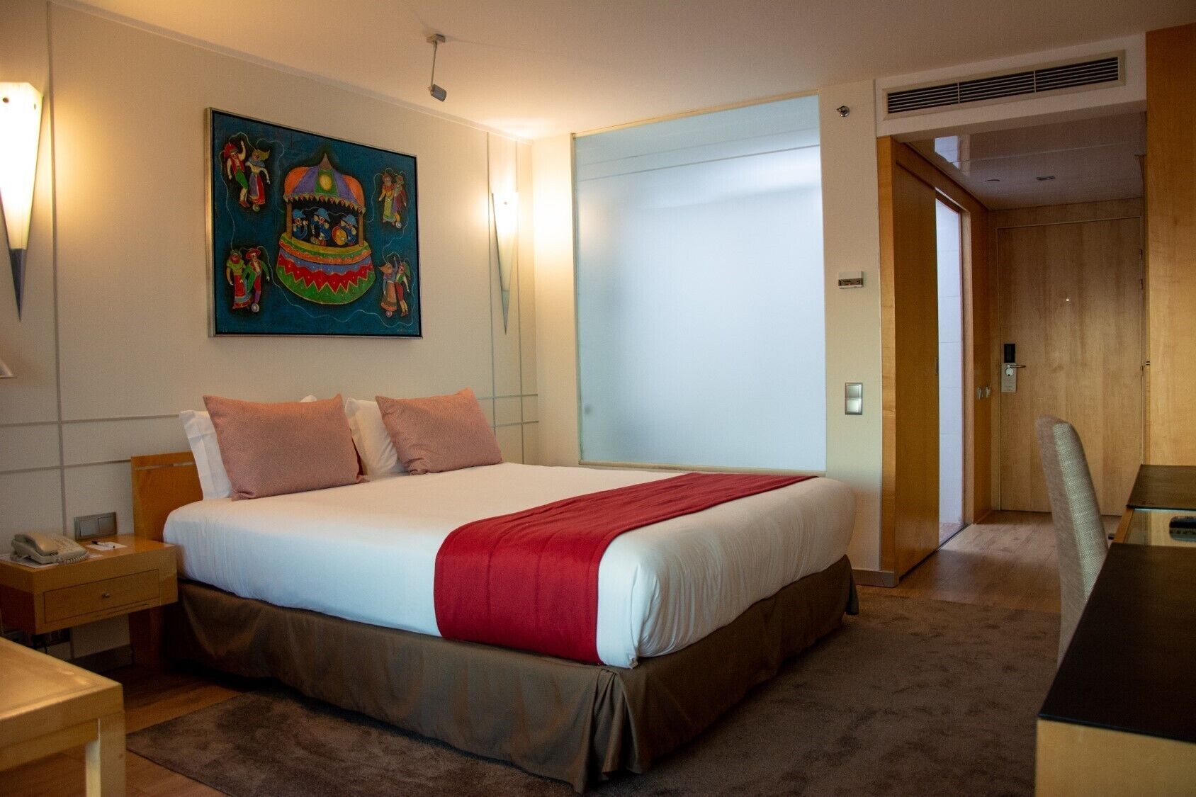 A hotel room with a bed, a desk, a chair, a painting on the wall, and a door leading to another room.