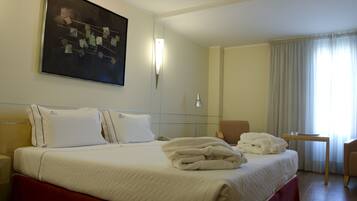 Una habitación de hotel con una cama bien hecha, una mesita de noche, una silla, un escritorio y un cuadro en la pared.