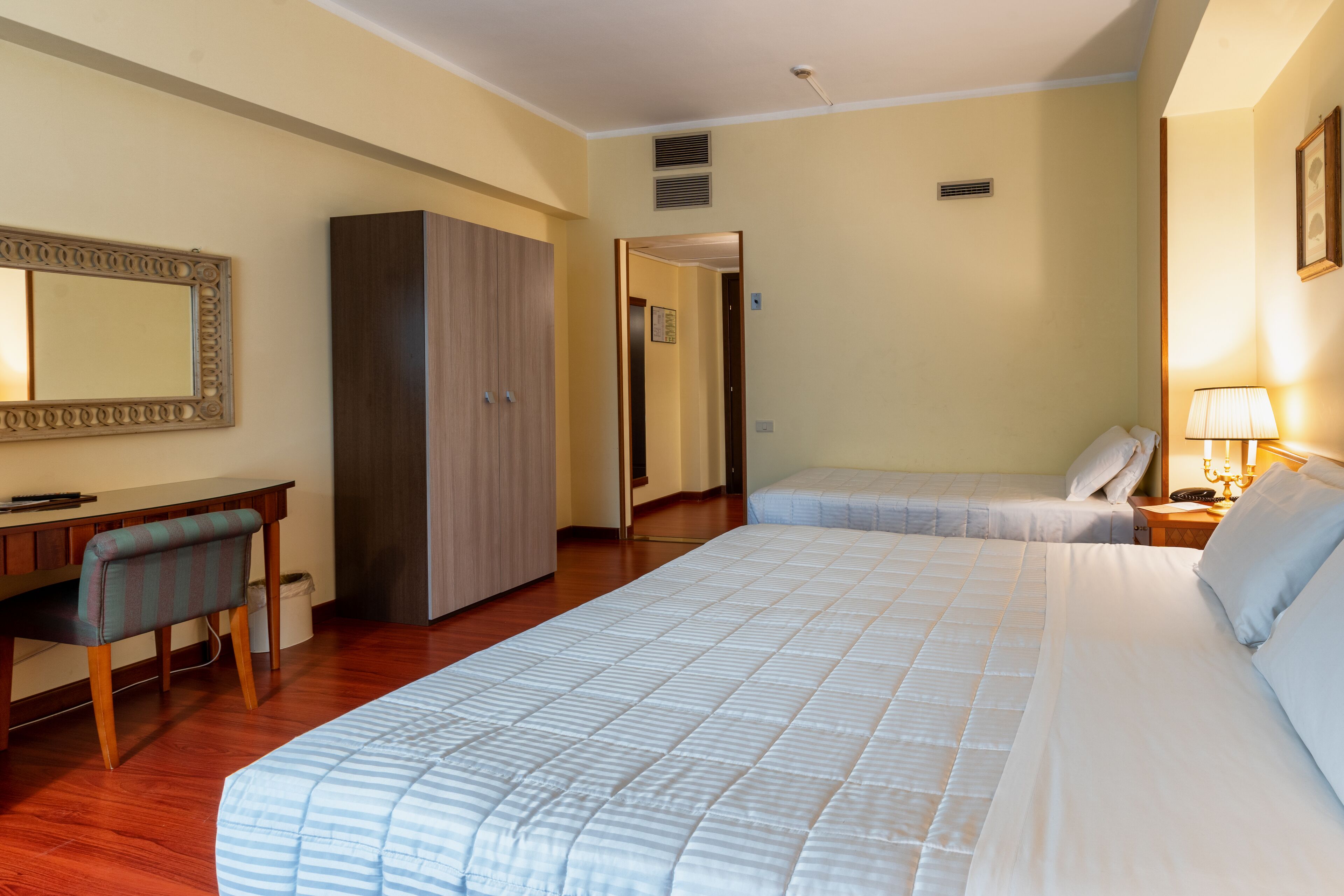 A hotel room with a large bed, a desk with a chair, a wardrobe, and a bedside table with a lamp.