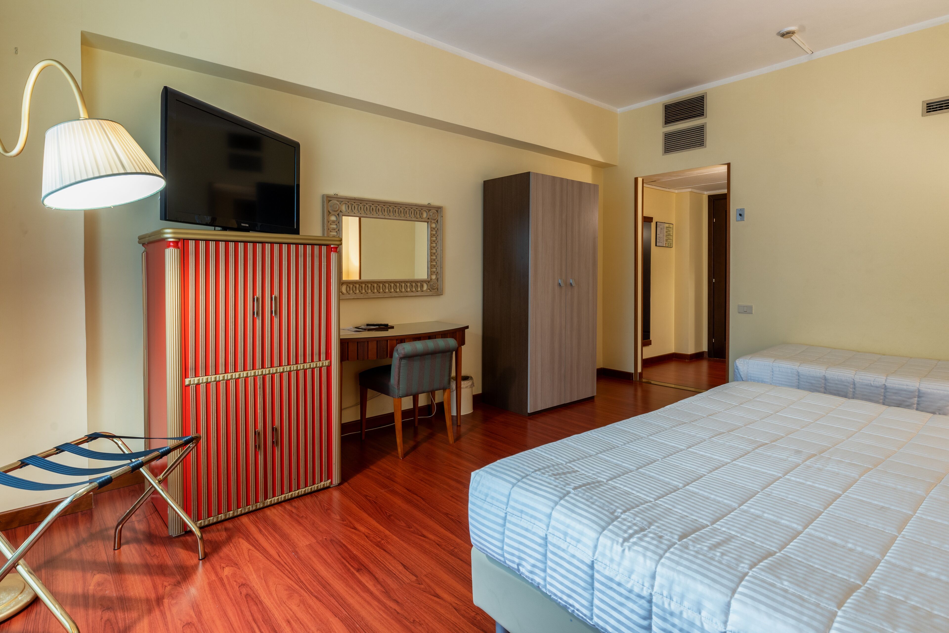 A hotel room with a bed, a desk, a chair, a wardrobe, a television, and a lamp.