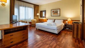 A hotel room with a wooden desk, a bed with white linens, a chair, a TV, and a lamp.