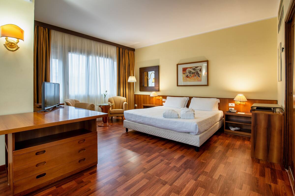 Une chambre d’hôtel avec un bureau en bois, un lit avec du linge de lit blanc, une chaise, un téléviseur et une lampe.