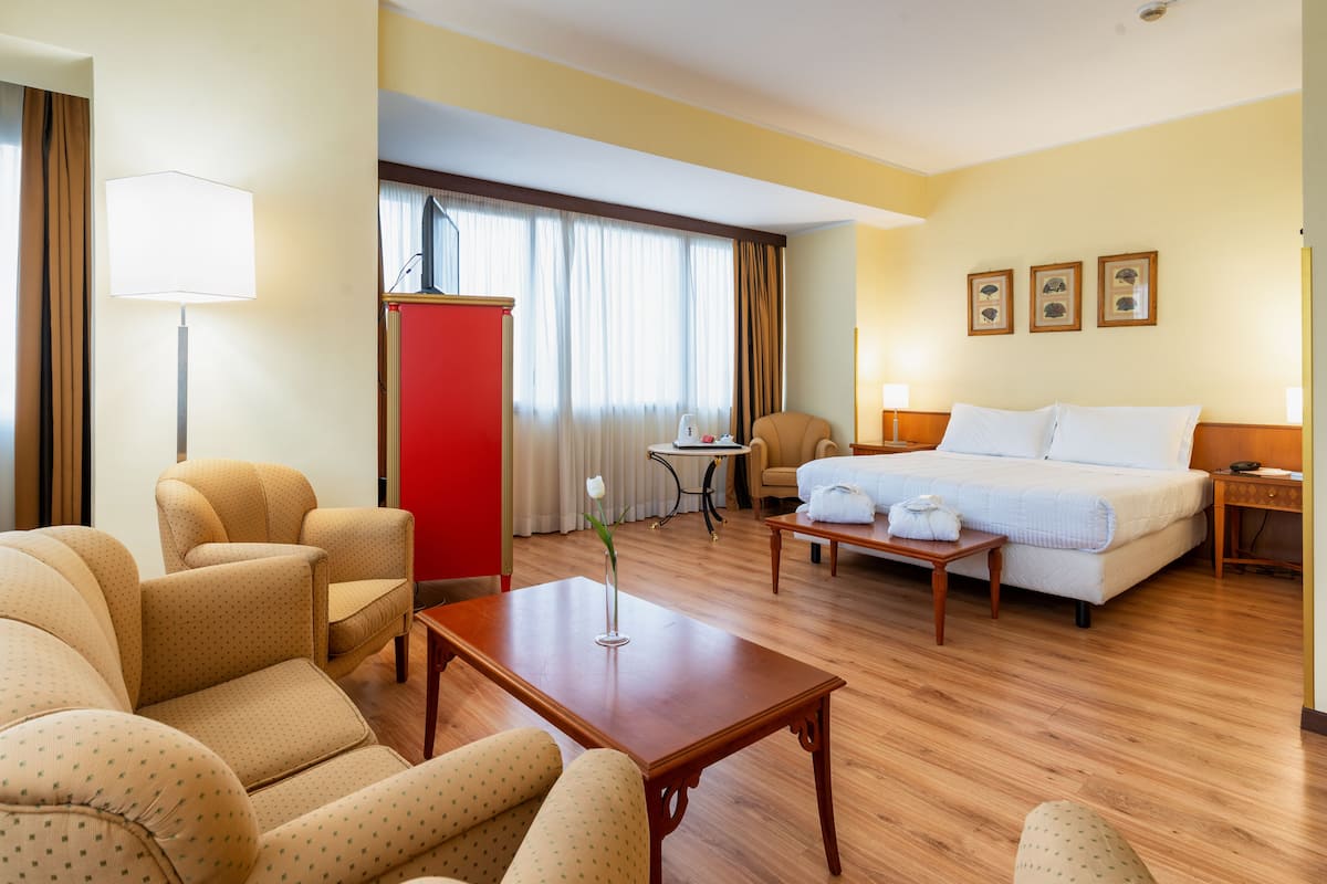 Une chambre d’hôtel avec un lit, deux fauteuils, une petite table, un bureau et un meuble rouge.