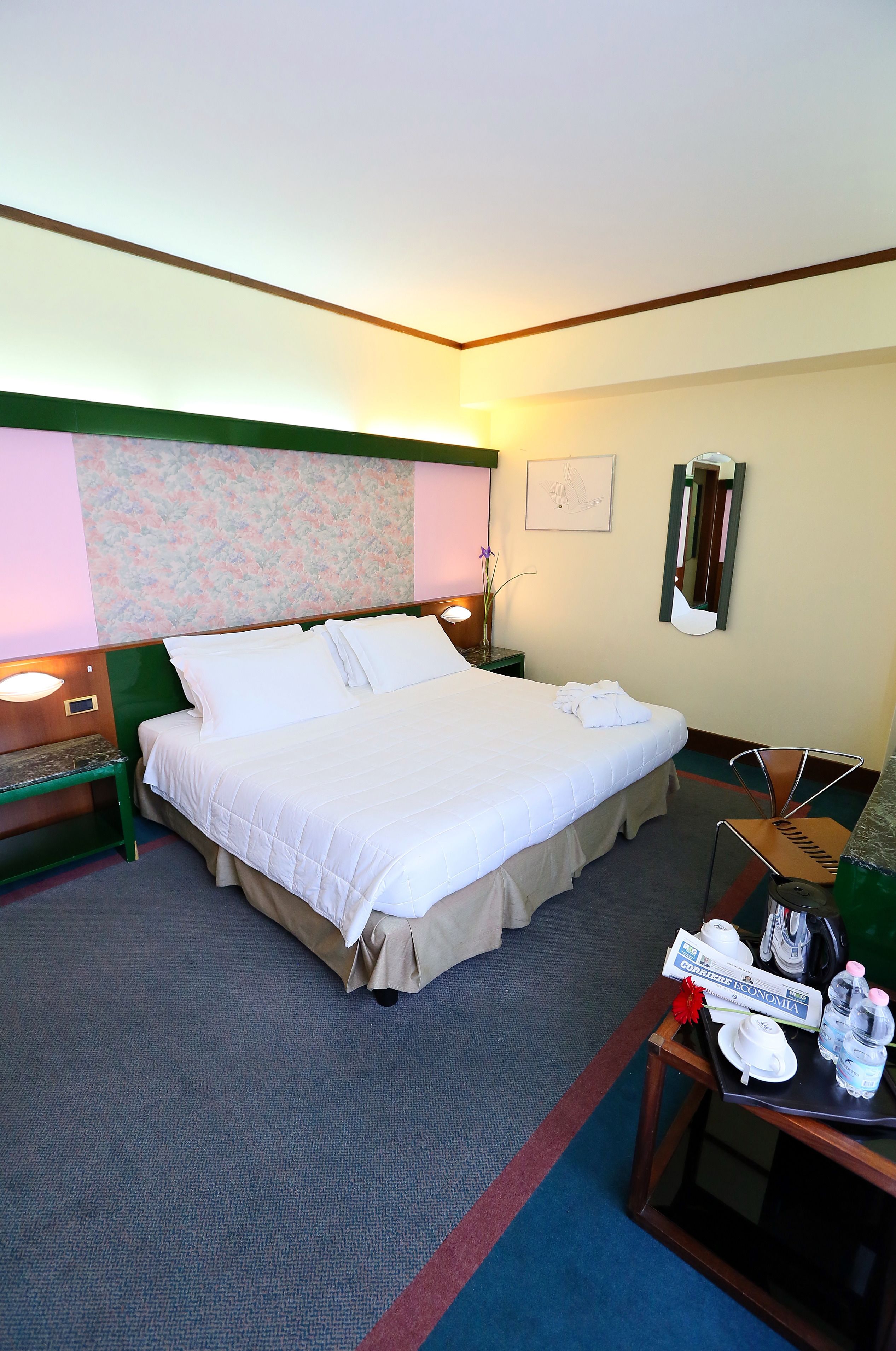 A hotel room with a large bed, a bedside table with a lamp, a chair, and a small table with a sign and some items on it.