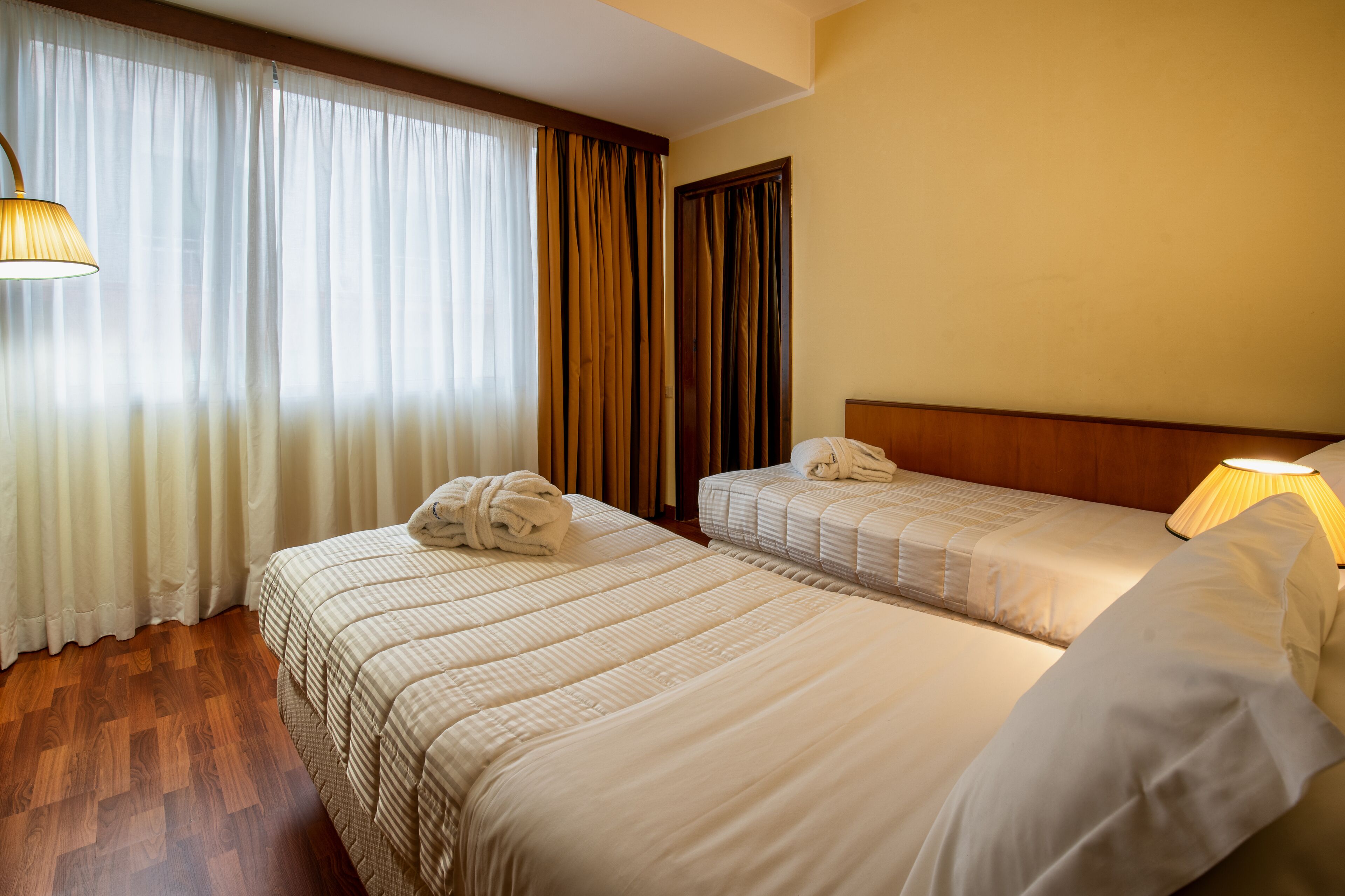 Habitación de hotel con dos camas, cabecero de madera, ventana con cortinas traslúcidas y una lámpara.