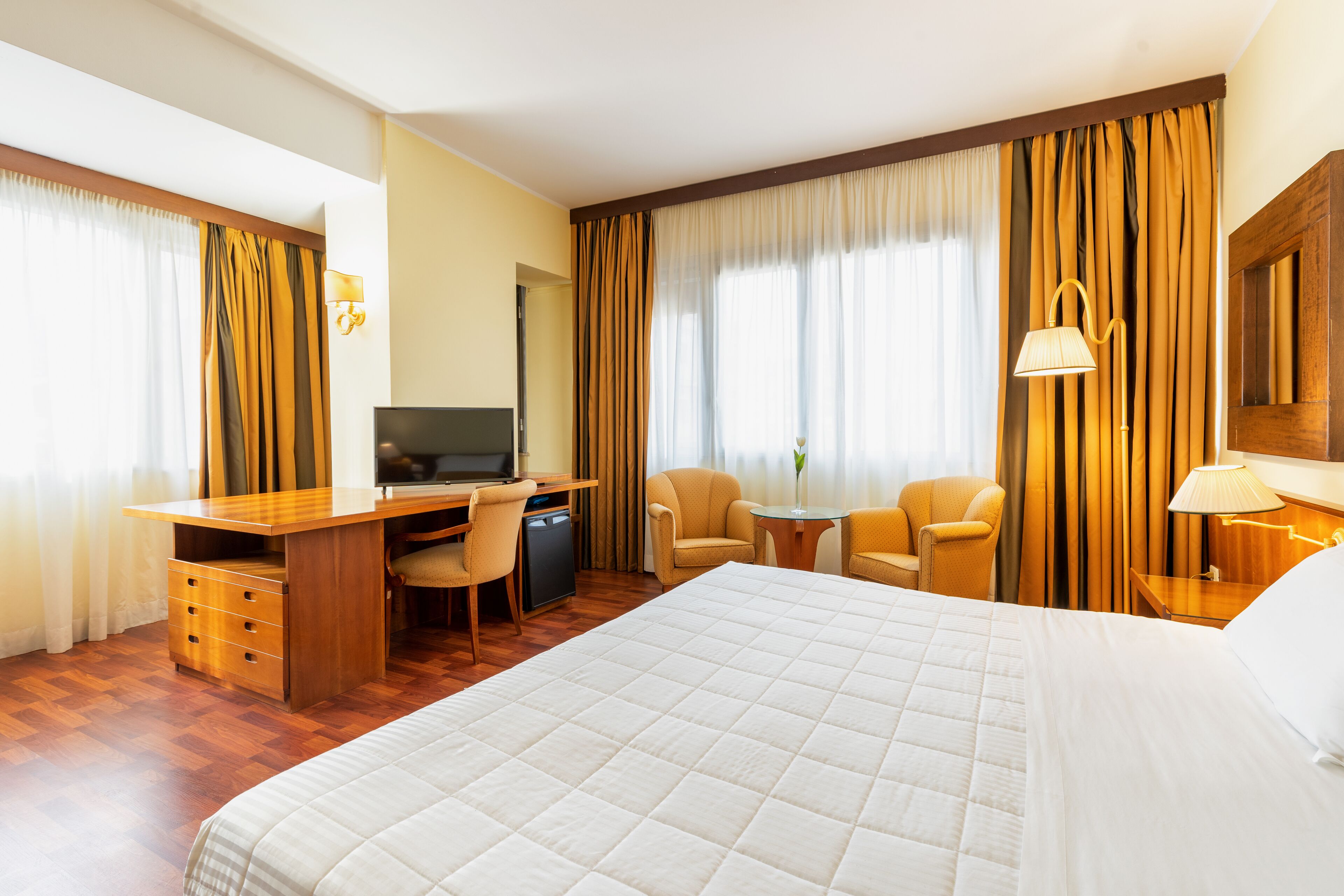 Una camera d'albergo con un letto grande, una scrivania con una sedia, una televisione e un tavolino con una lampada.